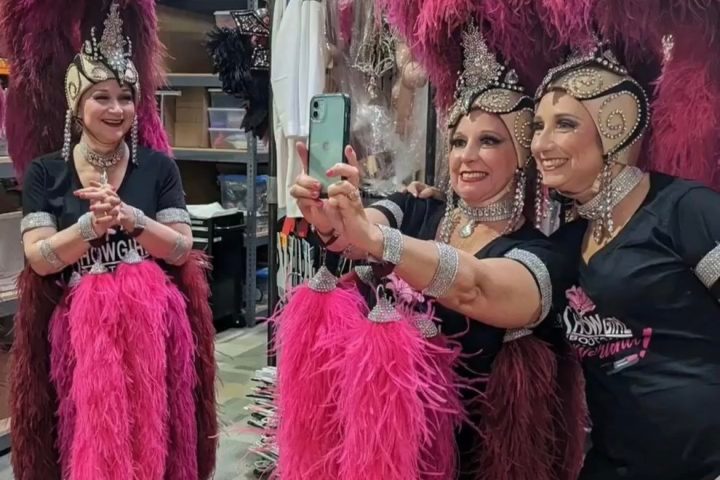 Three women in feathered showgirl costumes taking a mirror selfie, smiling in a dressing room.