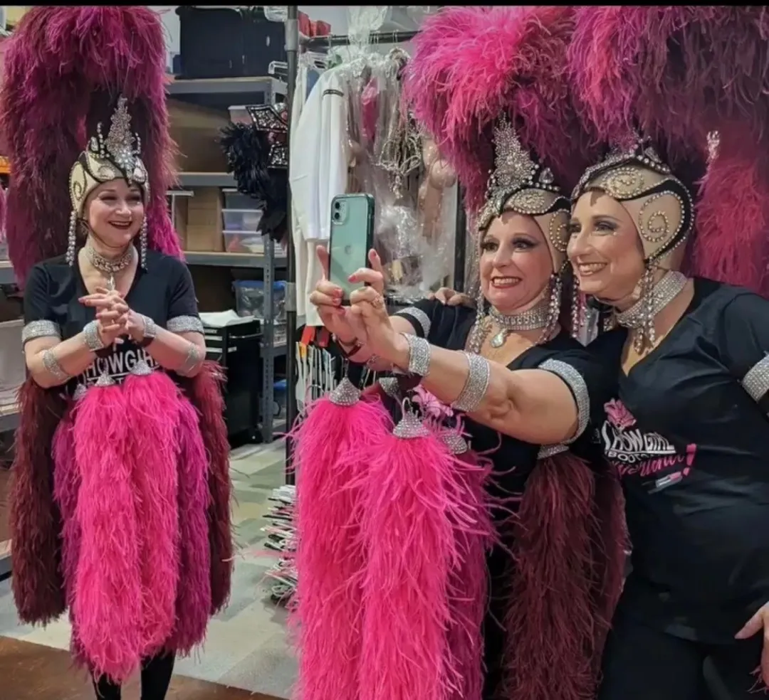 Three women in feathered showgirl costumes taking a mirror selfie, smiling in a dressing room.